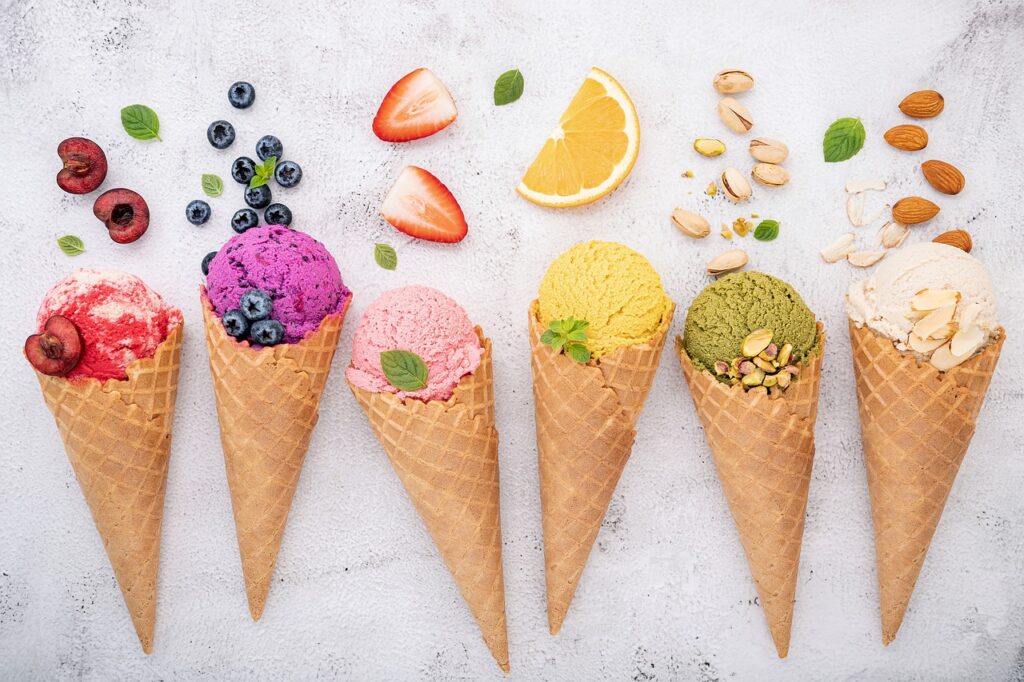 Picture of 6 ice cream cones laying on a counter.  All different colors with pieces of fruit above each cone.